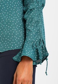 Türkise Bluse mit weißen Punkten, leichtes Material, geraffte Ärmel mit Bändern und einem ausgestellten Ärmeldesign.