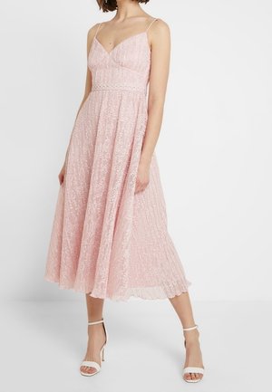 Kobieta w różowej, plisowanej sukience midi na ramiączkach, ubrana w białe sandały na wysokim obcasie, stojąca na jednolitym tle.