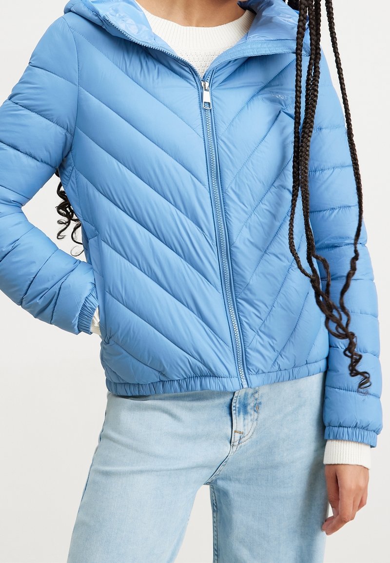 Prošívaná modrá bunda s kapucí, zdobená vzorem ve tvaru šipky, se zipem vpředu a elastickými manžetami. Natažená přes bílý pletený svetr.