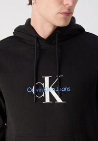Felpa nera con cappuccio e tasca a marsupio, caratterizzata da un grande logo bianco "CK" e dalla scritta "Calvin Klein Jeans" in blu. Realizzata in tessuto misto cotone.