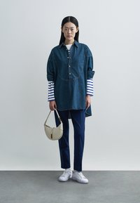 Sukienka koszulowa z denimu z przodem na guziki, podwiniętymi rękawami i zaokrąglonym dołem; zestawiona z granatowymi spodniami i białymi trampkami; trzyma beżową torebkę.