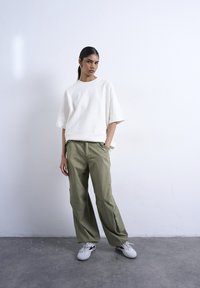 Νεαρή γυναίκα στέκεται μπροστά σε λευκό τοίχο, φορώντας υπερμεγέθη λευκό μπλουζάκι, φαρδιά πράσινα παντελόνια cargo και λευκά αθλητικά παπούτσια με μαύρες λεπτομέρειες.