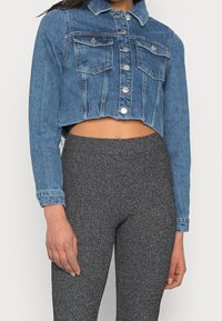 Krótkia kurtka dżinsowa w średnim odcieniu niebieskiego z przednimi kieszeniami, srebrnymi elementami i klasycznym kołnierzem. W zestawie z teksturowanymi szarymi leginsami.