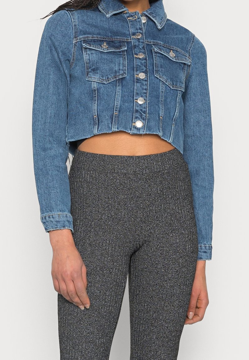 Krótkia kurtka dżinsowa w średnim odcieniu niebieskiego z przednimi kieszeniami, srebrnymi elementami i klasycznym kołnierzem. W zestawie z teksturowanymi szarymi leginsami.