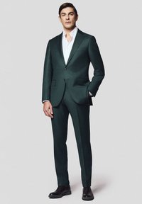 Ciemnozielony garnitur o dopasowanym kroju, biały podkoszulek, gładka tekstura i klasyczny kołnierz. Brązowe skórzane buty dopełniają całość.