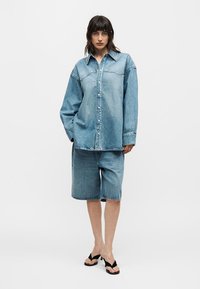 Γυναίκα που φοράει υπερμεγέθη ανοιχτό μπλε πουκάμισο από τζιν, τζιν σορτς μέχρι το γόνατο σε ταιριαστό χρώμα και μαύρα σανδάλια με τακούνι, στέκεται μπροστά σε απλό φόντο.