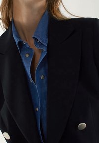 Veste noire croisée sur une chemise en denim bleu, avec des boutons argentés et une texture lisse, dotée de revers bien définis.