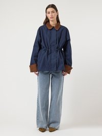 Γυναίκα που φοράει μπλε σκούρο μπουφάν με καφέ γιακά και μανσέτες, ανοιχτό μπλε τζιν παντελόνι με φαρδιά μπατζάκια και καφέ μυτερές μπότες, στέκεται μπροστά σε απλό φόντο.