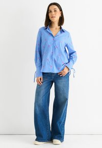 Blau gestreiftes Langarmhemd mit pinken Verzierungen; hat einen Kragen und verstellbare Ärmelbindungen. Kombiniert mit weit geschnittenen blauen Jeans.