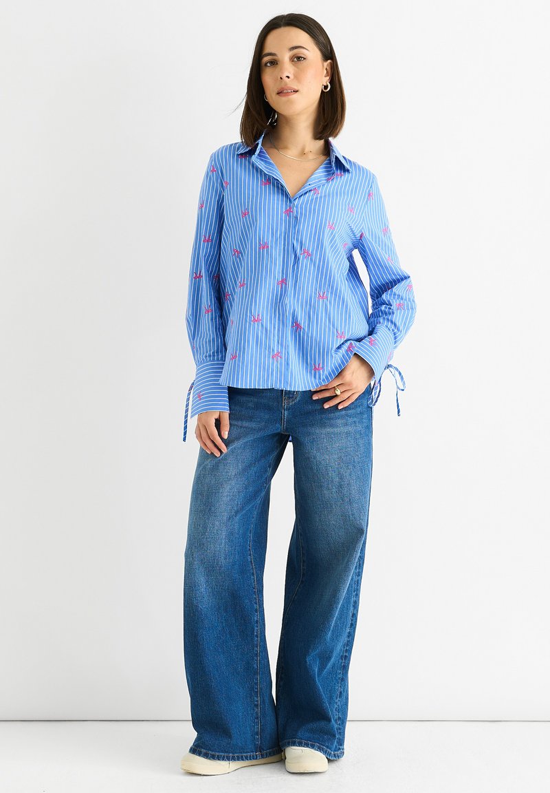 Blau gestreiftes Langarmhemd mit pinken Verzierungen; hat einen Kragen und verstellbare Ärmelbindungen. Kombiniert mit weit geschnittenen blauen Jeans.
