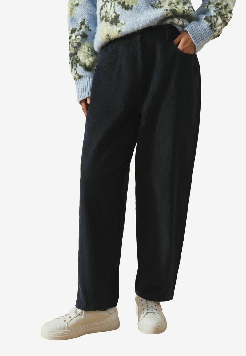 Tmavé kordové nohavice vo voľnom strihu s bočnými vreckami a hladkou textúrou, doplnené o biele tenisky. Sveter s kvetinovým vzorom.