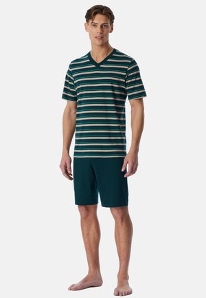 Mann steht barfuß, trägt ein grün-beige gestreiftes kurzärmliges V-Ausschnitt-Shirt und einfarbige grüne knielange Shorts vor einfarbigem Hintergrund.