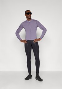 Camiseta deportiva de manga larga morada con cremallera frontal, combinada con leggings negros ajustados. La modelo lleva zapatos negros y gafas de sol.