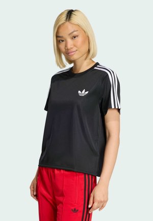 Giovane donna con capelli biondi lisci che indossa una maglietta nera Adidas con strisce bianche e pantaloni rossi Adidas con strisce nere.