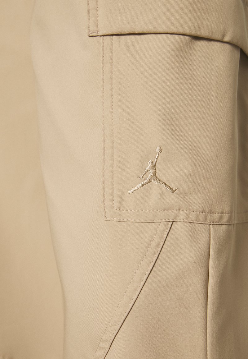 Pantaloni cargo beige realizzati in tessuto liscio, caratterizzati da una tasca cucita prominente e da un logo Jumpman ricamato in filo dorato.