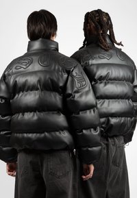 Black puffer jacket made of smooth, shiny material featuring large, embossed lettering on the back, with adjustable hem and relaxed fit.