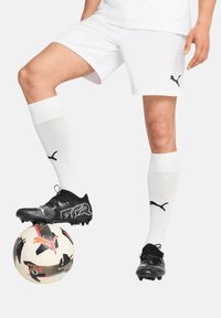 Schwarze Fußballschuhe mit strukturierter Oberfläche, weiße kniehohe Socken, weiße Shorts und ein gemusterter Fußball unter dem Fuß.
