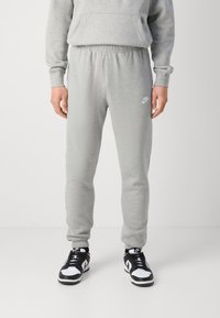 Osoba oblečená v šedých teplácích Nike a černo-bílých teniskách Nike, stojící na jednobarevném bílém pozadí.