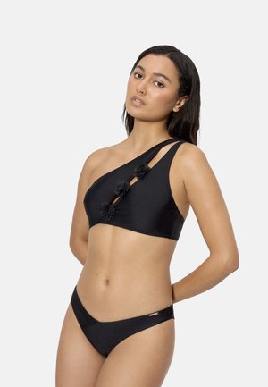 Mujer con top de bikini negro de un solo hombro con cortes florales y braguitas bajas a juego, de pie contra un fondo liso.