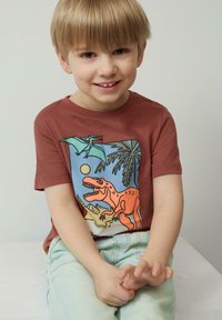 Kortebloemige roestkleurige T-shirt met een kleurrijke dinosaurustekening. Lichtblauwe spijkerbroek. Zachte stof, ontspannen pasvorm, casual ontwerp.
