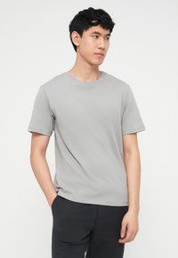 Νέος άνδρας με μαύρα μαλλιά, που φοράει ανοιχτογκρι κοντομάνικο μπλουζάκι και σκουρόχρωμο παντελόνι, κοιτάζει προς τα δεξιά του με το ένα χέρι στη ν τσέπη.