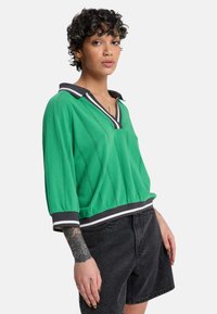 Pull-over vert en maille avec col en V, col contrastant noir et blanc, et rayures texturées. Doté de manches amples et d'une coupe courte.