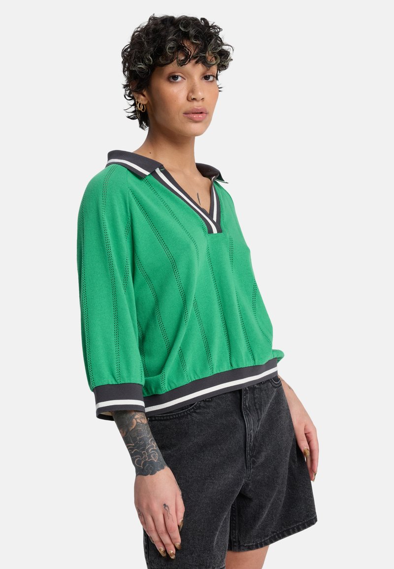 Pull-over vert en maille avec col en V, col contrastant noir et blanc, et rayures texturées. Doté de manches amples et d'une coupe courte.