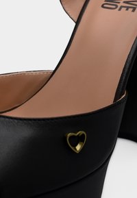 Κοντινή λήψη μαύρου παπουτσιού με ψηλά τακούνια, με ταμπά εσωτερικό και μικρή μεταλλική χρυσή διακόσμηση σε σχήμα καρδιάς στο πλάι.