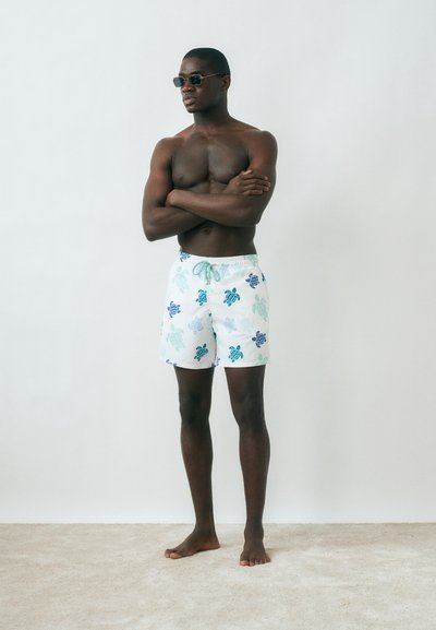 Μυώδης άνδρας που φοράει μαγιό με σχέδιο χελώνας και γυαλιά ηλίου, στέκεται ξυπόλητος με τα χέρια σταυρωμένα μπροστά σε απλό λευκό φόντο.