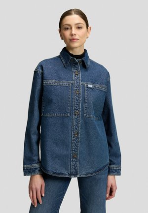Junge Frau trägt eine dunkelblaue Jeansjacke über passenden Jeans und einem schwarzen Top, steht vor einem einfarbigen hellgrauen Hintergrund.