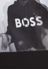 Felpa nera con cappuccio caratterizzata da un grande testo bianco "BOSS" con un ritaglio circolare nella lettera "O". Tessuto morbido, design informale.
