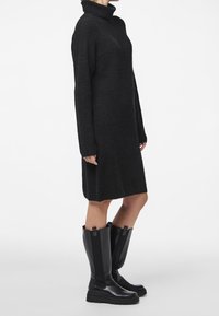 Robe en maille noire avec un col haut, des manches longues et une coupe décontractée. Portée avec des bottes noires hautes à talon léger et finition texturée.