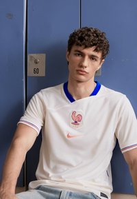 Biała koszulka sportowa wykonana z oddychającego materiału, z emblematem koguta, logo Nike oraz niebieskimi i czerwonymi akcentami na kołnierzyku i rękawach.