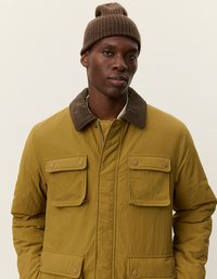 Gulgrønn isolert jakke med to frontlommer med klaff, brun kordfløyelskrage og matt finish, båret med en brun strikket lue.