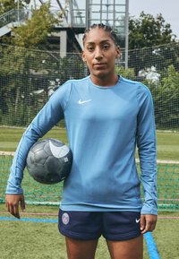 Nike hosszú ujjú kék póló kerek nyakkal, sötét rövidnadrággal, amelyen Párizs-logó látható. Egy fekete focilabdát tart, fehér részletekkel.