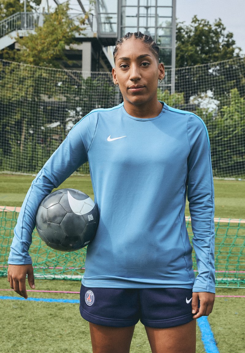 Nike hosszú ujjú kék póló kerek nyakkal, sötét rövidnadrággal, amelyen Párizs-logó látható. Egy fekete focilabdát tart, fehér részletekkel.