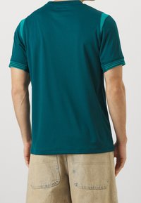 Camiseta deportiva color verde azulado con cuello redondo, mangas cortas y detalles a rayas en los hombros, combinada con pantalones cortos beige que muestran bolsillos traseros.