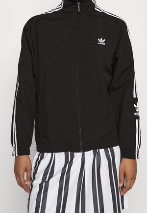 Veste de survêtement Adidas noire avec des bandes blanches sur les manches et un logo sur la poitrine et le bras, portée sur un pantalon à rayures verticales noir et blanc.