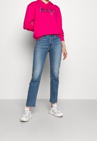 Fuchsiakleurige hoodie met trekkoorden en grafische tekst, gecombineerd met lichtblauwe jeans met rechte pijpen en witte sneakers. Zachte stof en een casual pasvorm.