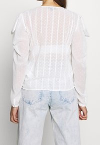 Blouse blanche transparente avec un motif texturé de points et de rayures verticales, dotée de manches bouffantes et d'un ourlet arrondi, portée avec un jean en denim clair.