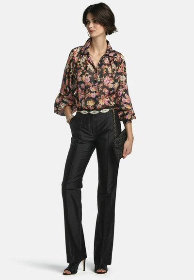 Donna che indossa una camicetta a maniche lunghe a fiori, pantaloni neri a vita alta, tacchi neri aperti sulle dita, che tiene una pochette nera con tracolla a catena.