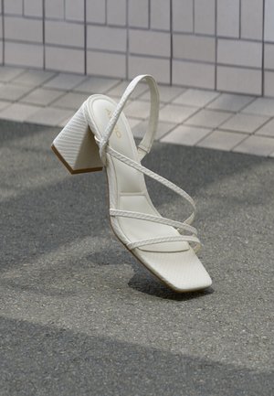 Witte sandaal met blokhak, gekruiste bandjes, gestructureerd bovenwerk en een puntige neus. Zachte binnenvoering en een gladde buitenzool.