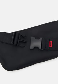 Marsupio in tessuto nero con una superficie liscia, dotato di fibbia a clip in plastica, chiusura a zip e un'etichetta logo rossa.