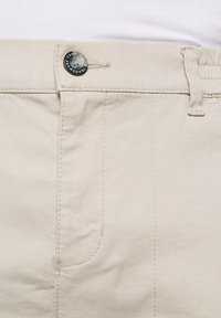 Pantalons en coton beige clair avec un tissu texturé, un bouton en métal avec un logo, et un design traditionnel à cinq poches.