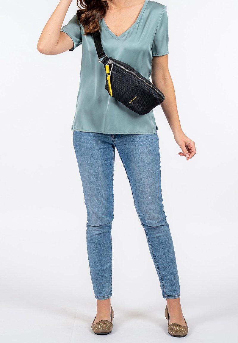 Borsa a tracolla nera con texture, dotata di una zip gialla, indossata sopra una blusa teal. Abbinata a jeans azzurri chiari e scarpe basse beige.