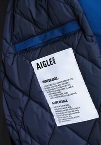 Intérieur d'une veste matelassée marine avec poche zippée bleue et étiquette blanche cousue arborant le logo AIGLE 1853 et un texte bilingue inspiré de la nature.