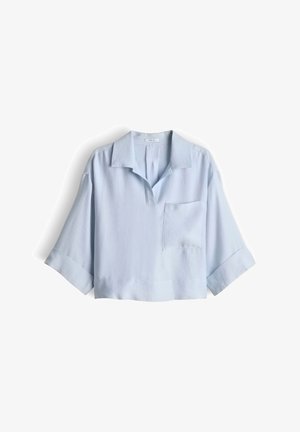 Chemise bleu clair à coupe ample avec de larges manches courtes, col ouvert et une unique poche poitrine côté droit, étalée à plat sur un fond blanc.