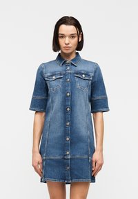 Γυναίκα που φοράει κοντομάνικο φόρεμα από τζιν με κουμπιά και τσέπες στο στήθος, στέκεται μπροστά σε απλό ανοιχτό φωτεινό φόντο.