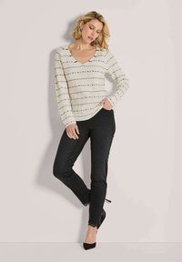 Frau lehnt an der Wand und trägt einen cremefarbenen V-Ausschnitt-Pullover mit schwarzen Streifen, schwarze enge Jeans mit Spitzenabschluss und schwarze High Heels.