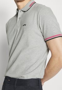 Grå kortärmad polo med en svart och röd randig krage, med en liten svart logotyp på bröstet. Slätt, lätt tyg.